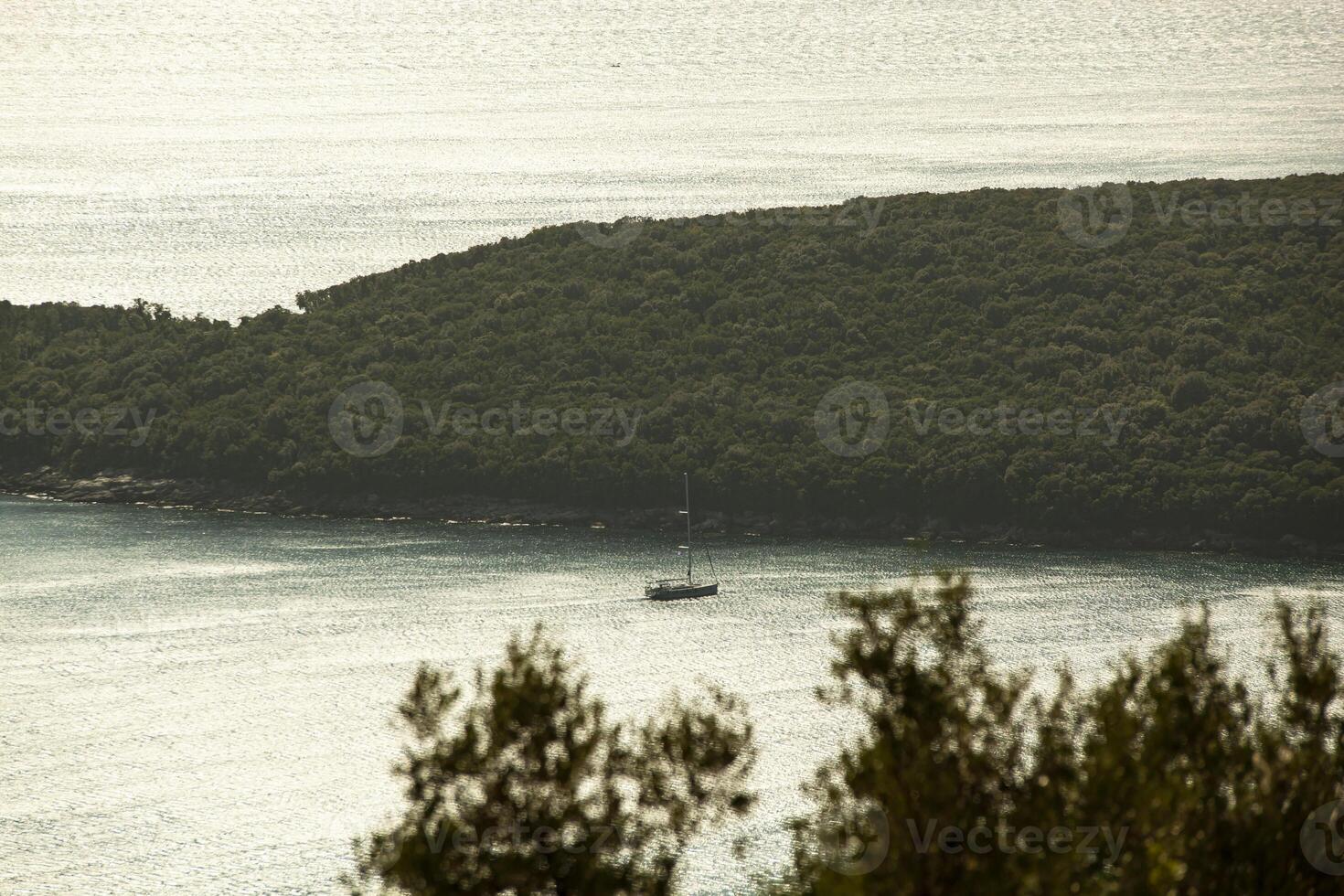 A small sailing ship sailing along the sea surface of the Adriatic Sea. An island with green trees. View from above. Montenegro. Budva. Horizontal. photo