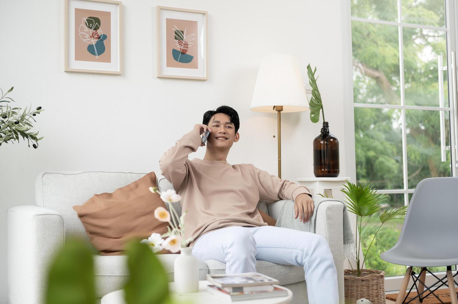 Happy asian man sitting on couch using smartphone in living room at home, relax time and lifestyle concept photo