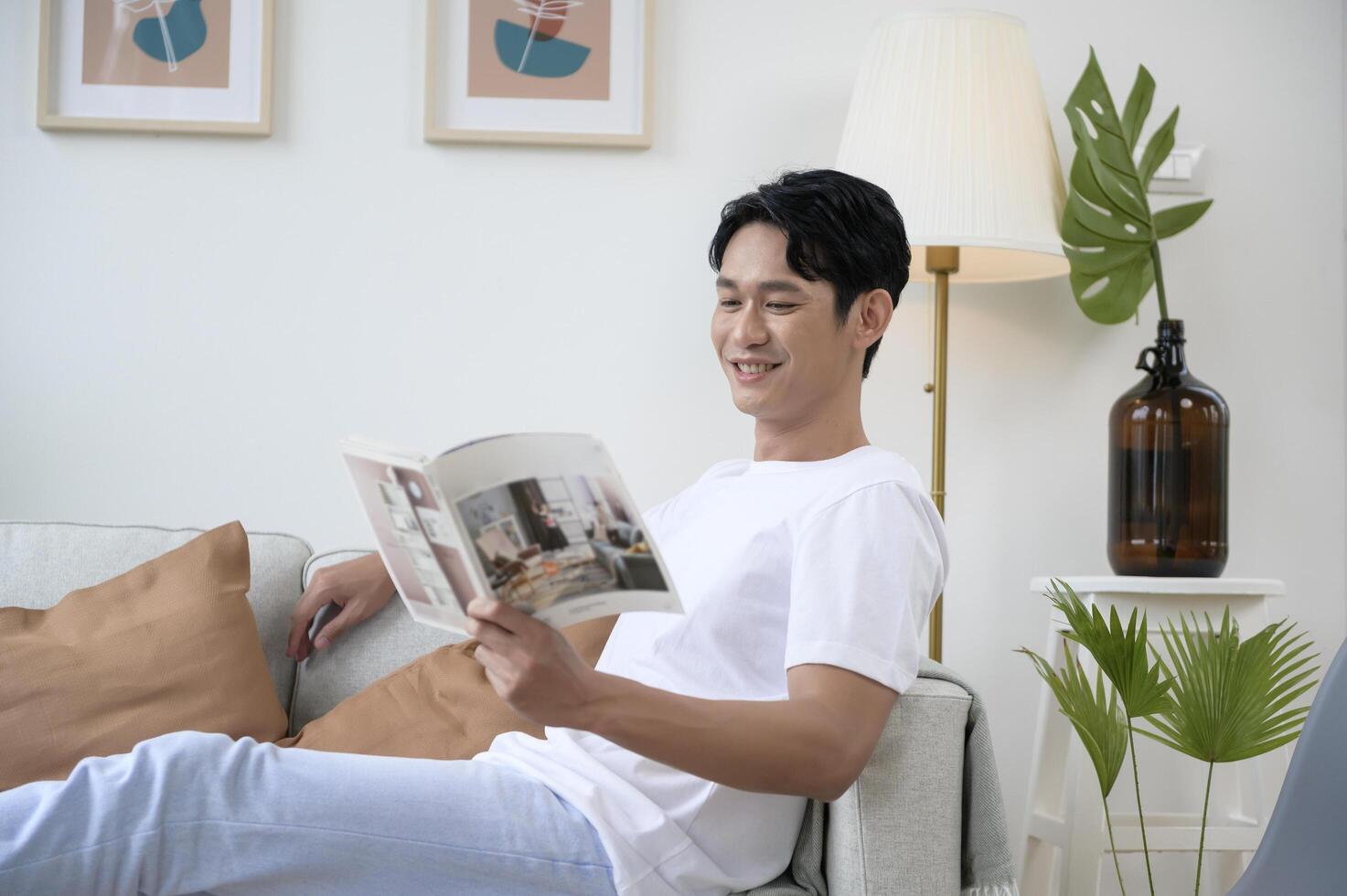 contento asiático hombre sentado en sofá leyendo un libro en vivo habitación a hogar, relajarse hora y estilo de vida concepto foto