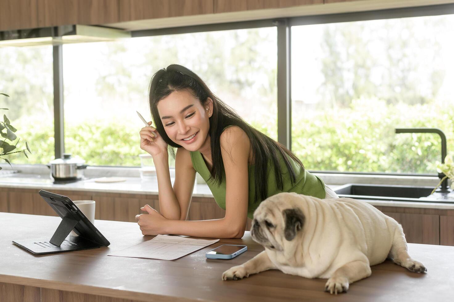joven asiático mujer trabajando con tableta y disfrutando con su perro en el cocina a hogar foto