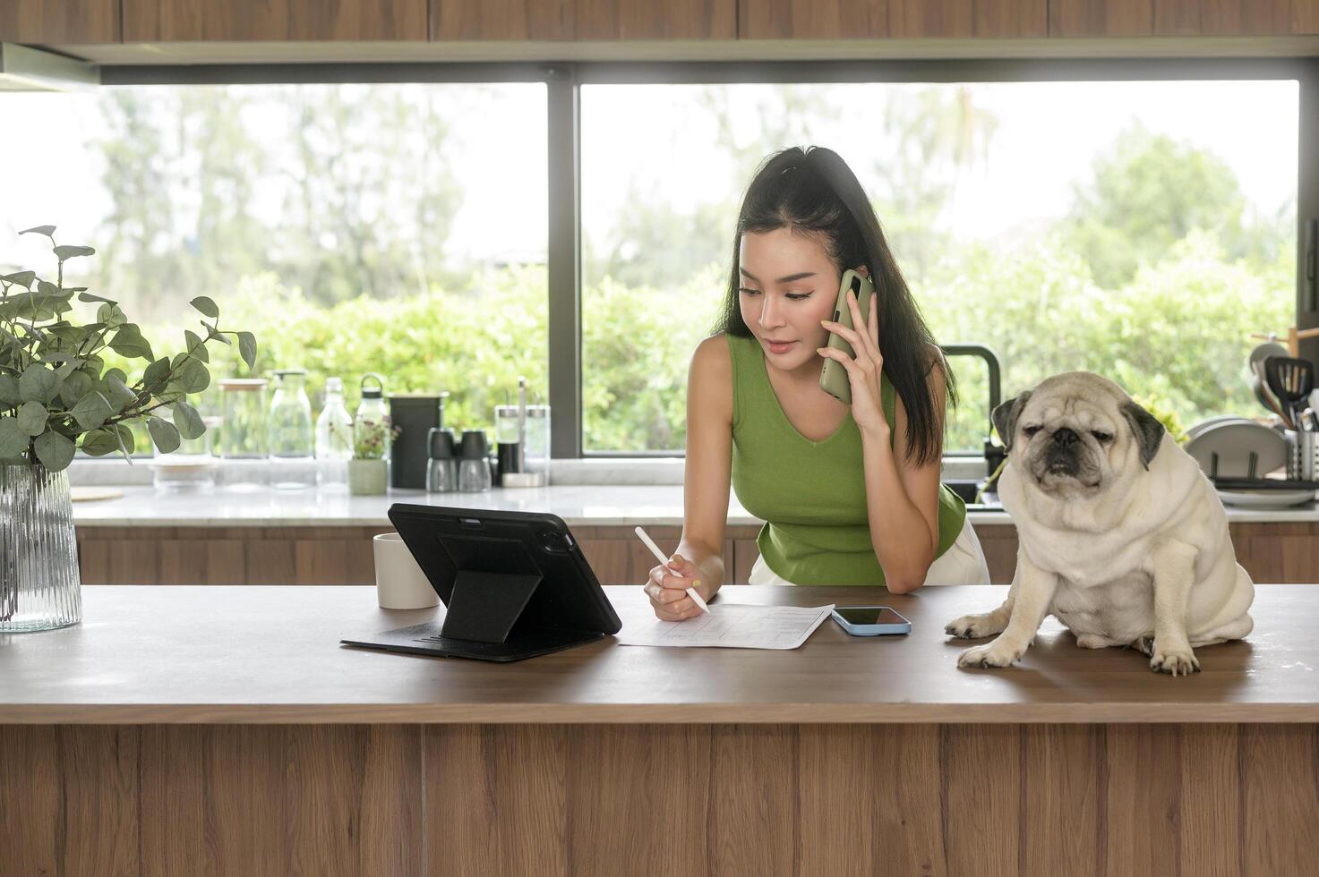 joven asiático mujer trabajando con tableta y disfrutando con su perro en el cocina a hogar foto