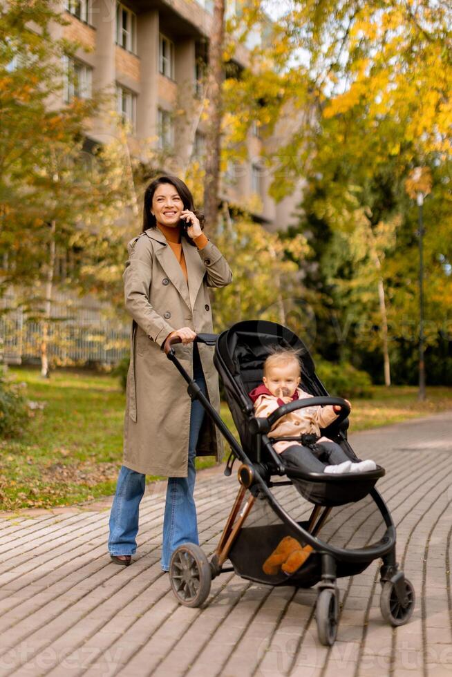 Young woman with cute baby girl in baby stroller using mobile phone at the autumn park photo