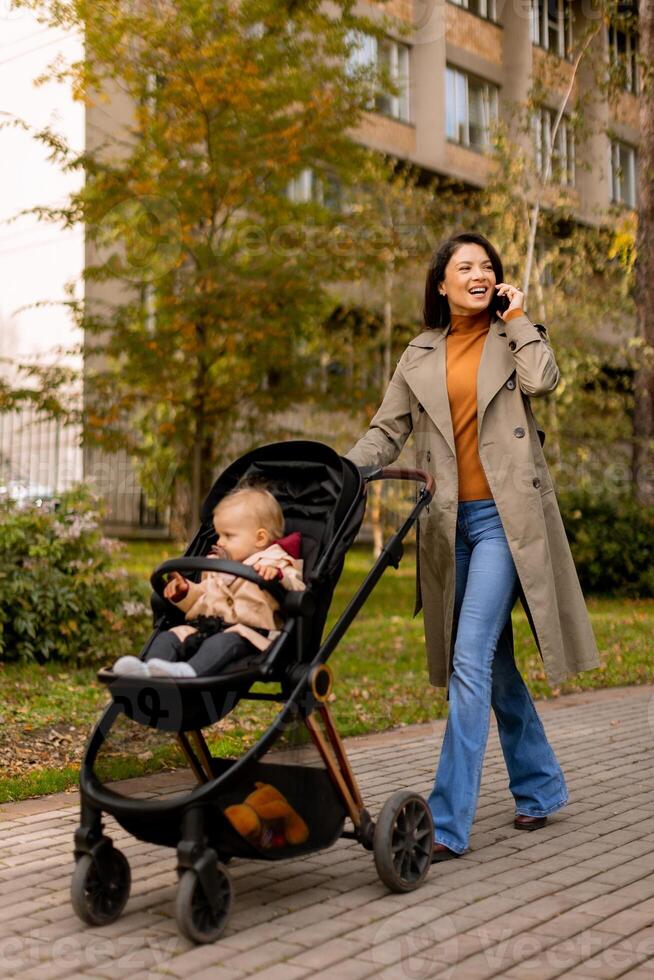 Young woman with cute baby girl in baby stroller using mobile phone at the autumn park photo