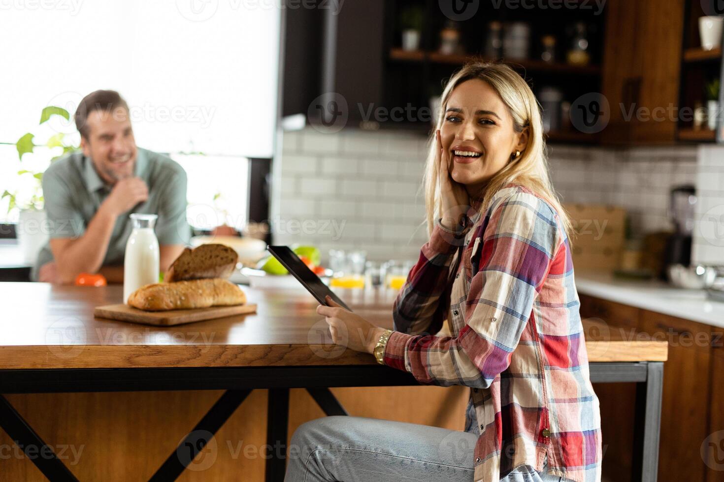 A woman reads from a tablet at the kitchen counter while a man holding an tomatoes smiles at her photo