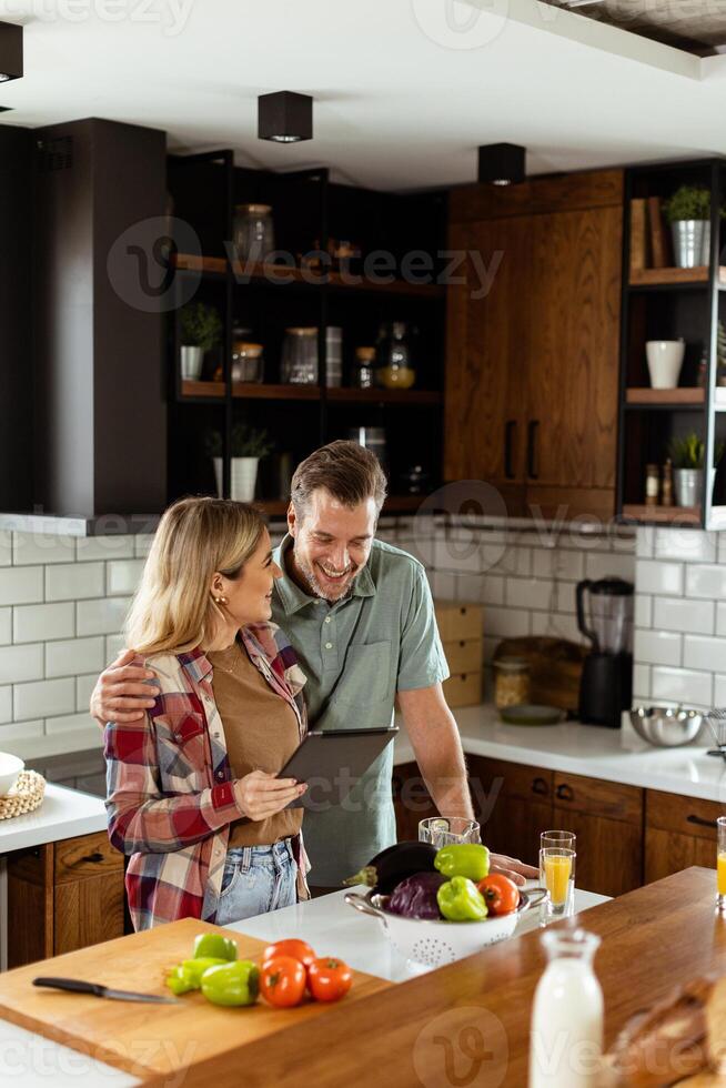 Cheerful couple stands in a well-lit kitchen, engrossed in a digital tablet among fresh ingredients photo