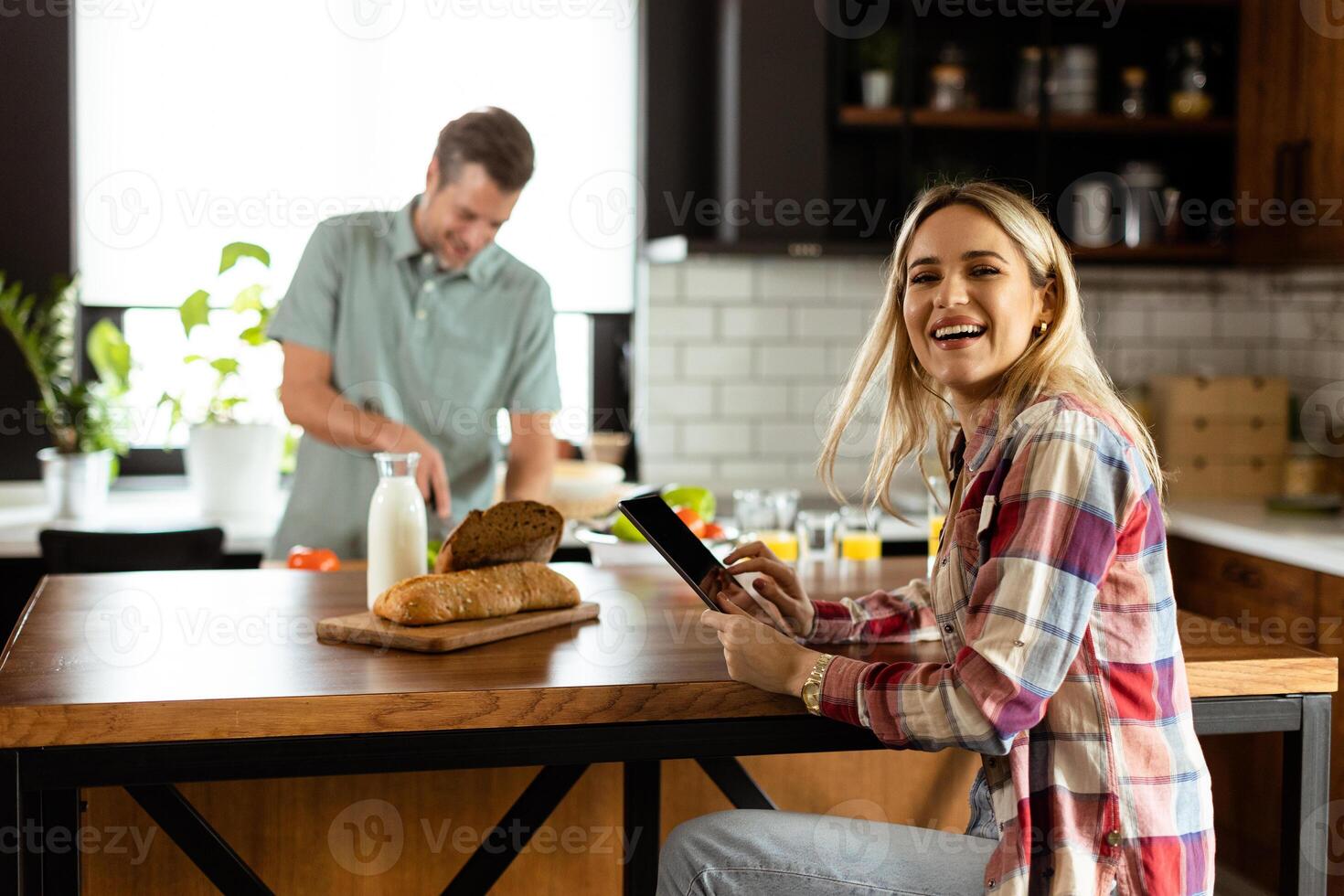 A woman reads from a tablet at the kitchen counter while a man holding an tomatoes smiles at her photo