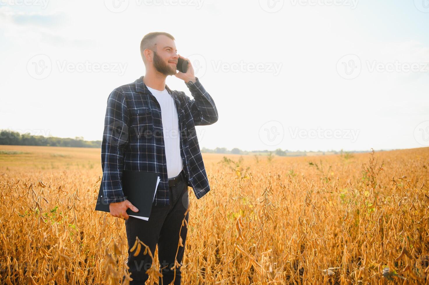 un linda granjero es en pie en un haba de soja campo y hablando en el teléfono con su negocio compañero foto