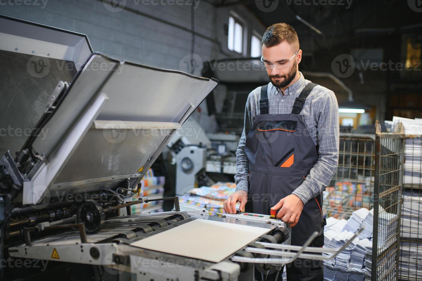 Printing house interior with modern offset print machine and operator in working uniform checking quality and controlling process of print photo