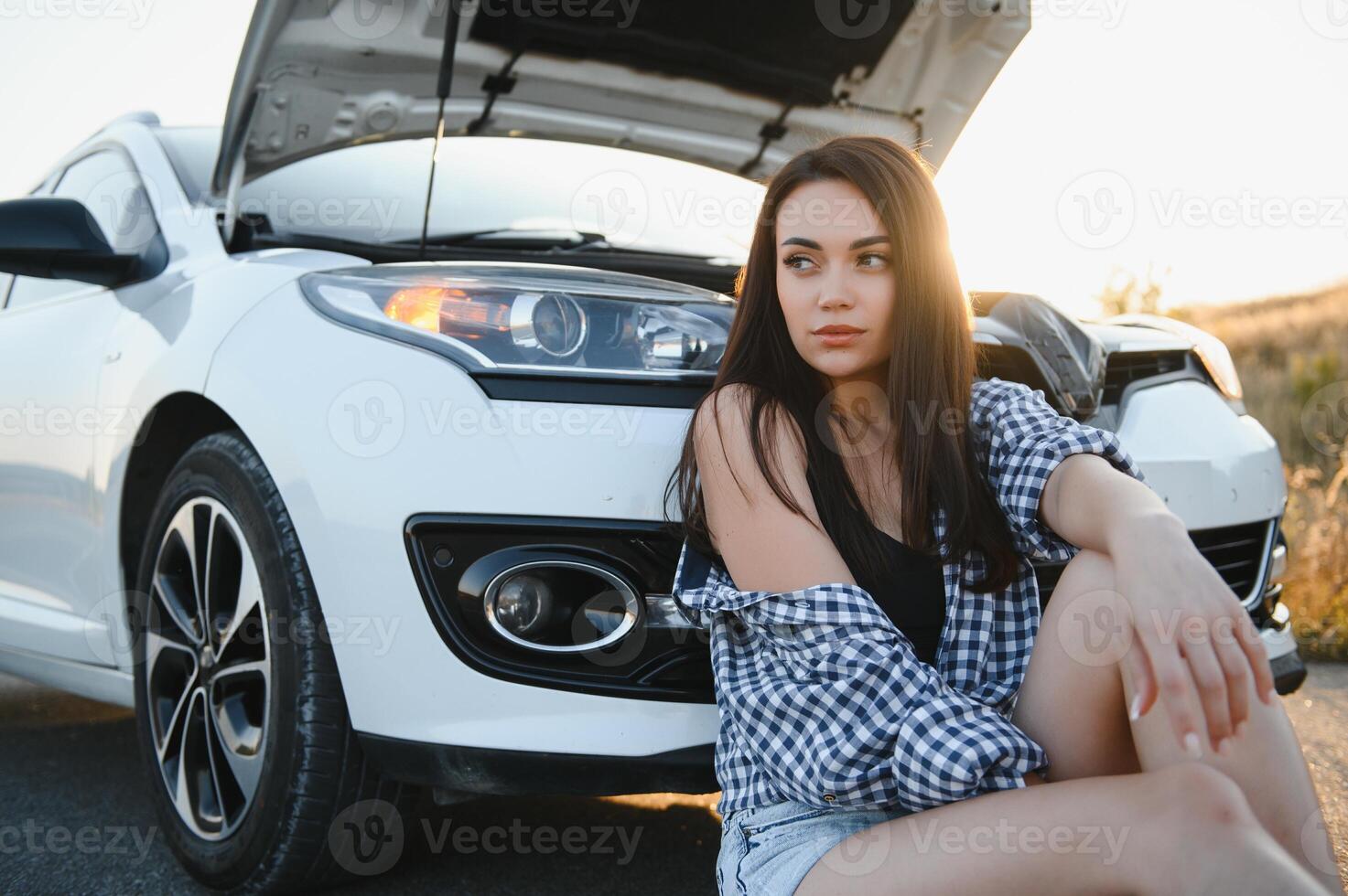 A young girl sits near a broken car on the road with an open hood. photo