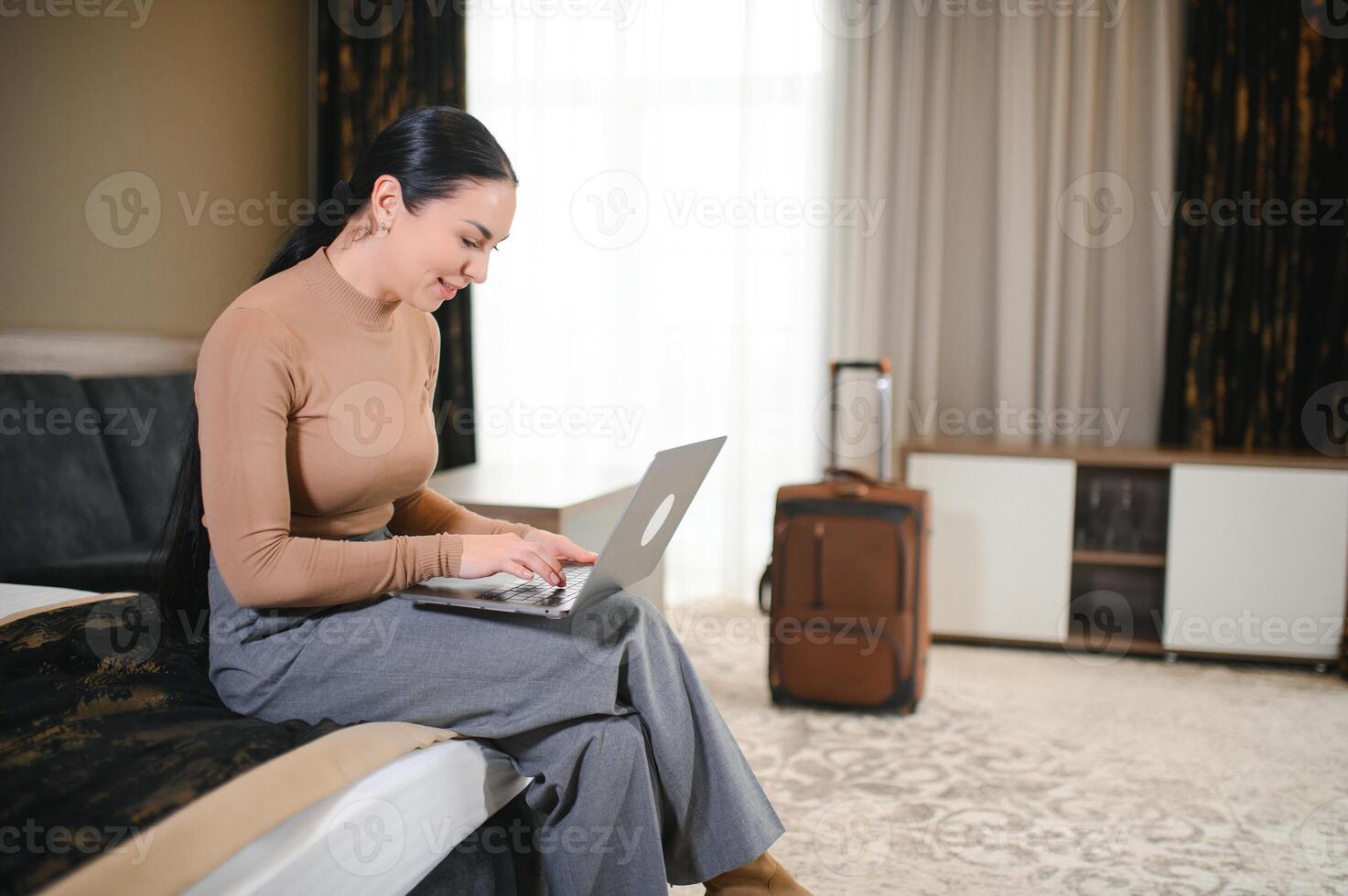 brunette white businesswoman sitting on bed in a hotel room talking on mobile phone using laptop computer photo