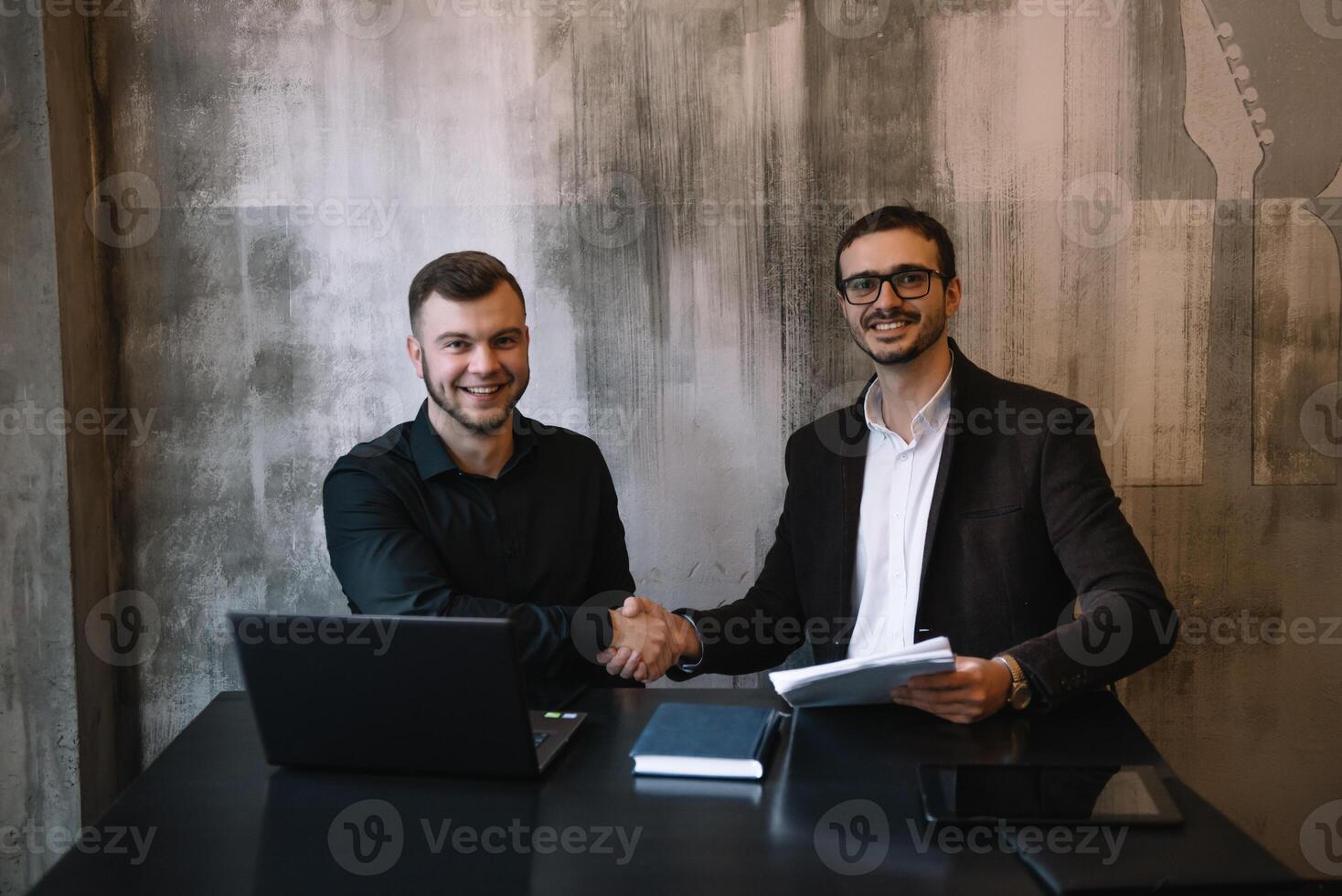 dos empresarios en un oficina sonriente a el cámara mientras trabajando juntos detrás un ordenador portátil computadora. foto