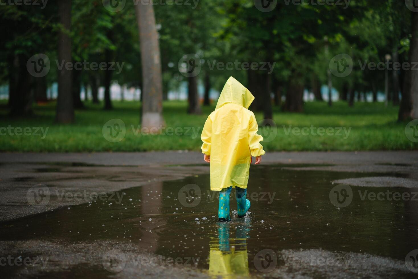 Little boy playing in rainy summer park. Child with umbrella, waterproof coat and boots jumping in puddle and mud in the rain. Kid walking in summer rain Outdoor fun by any weather. happy childhood. photo