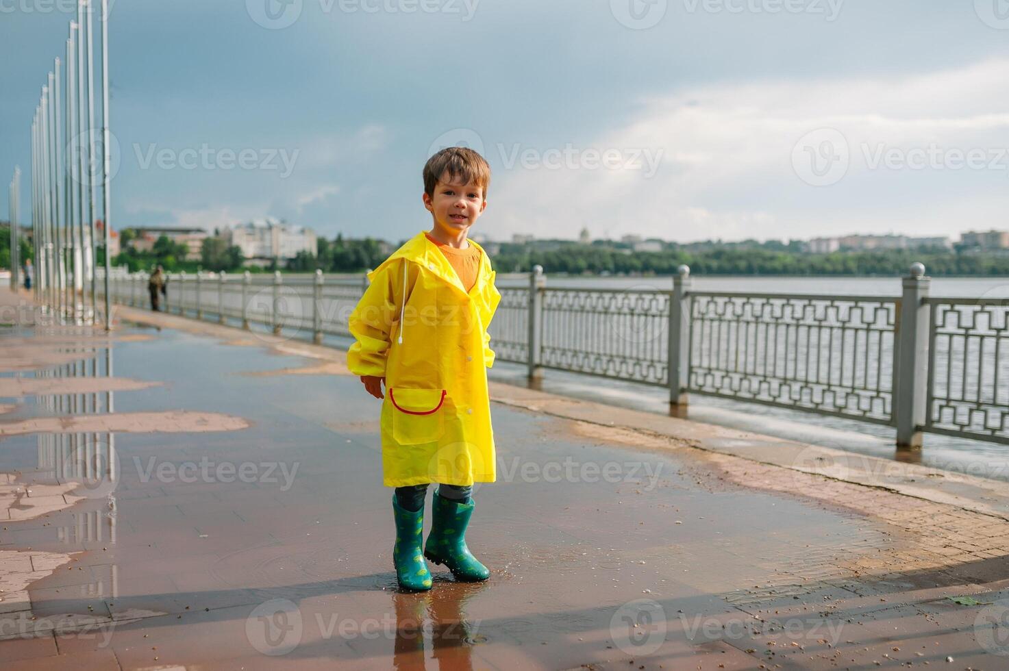 Little boy playing in rainy summer park. Child with umbrella, waterproof coat and boots jumping in puddle and mud in the rain. Kid walking in summer rain Outdoor fun by any weather. happy childhood. photo