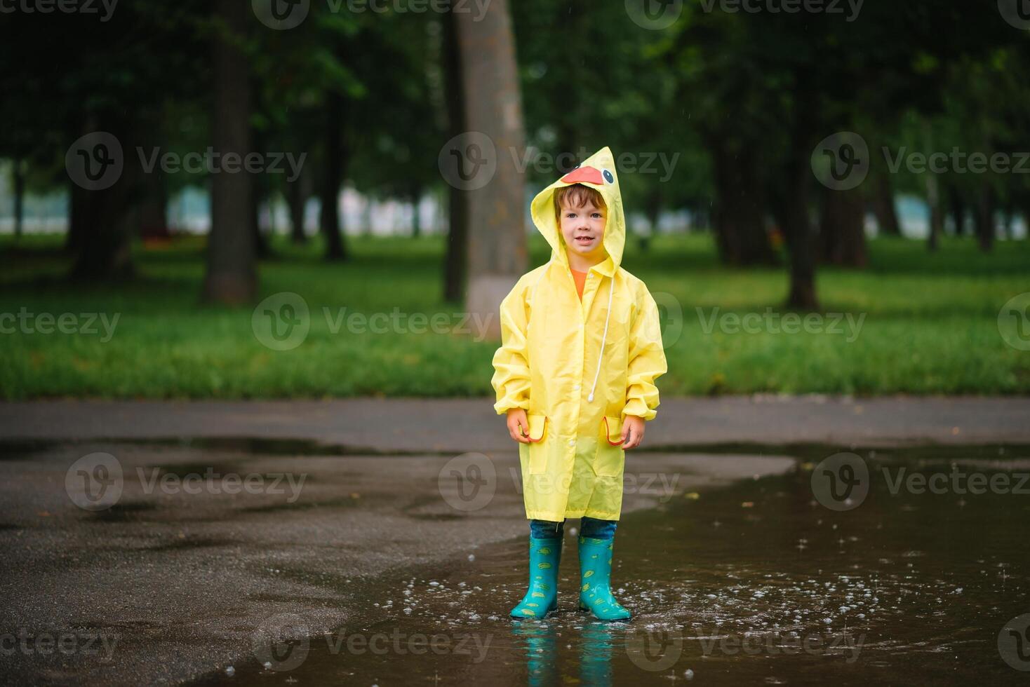 Little boy playing in rainy summer park. Child with umbrella, waterproof coat and boots jumping in puddle and mud in the rain. Kid walking in summer rain Outdoor fun by any weather. happy childhood. photo