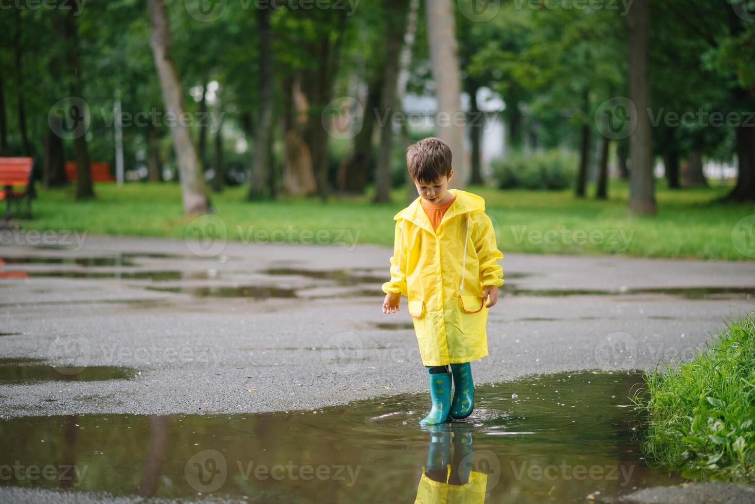 Little boy playing in rainy summer park. Child with umbrella, waterproof coat and boots jumping in puddle and mud in the rain. Kid walking in summer rain Outdoor fun by any weather. happy childhood photo