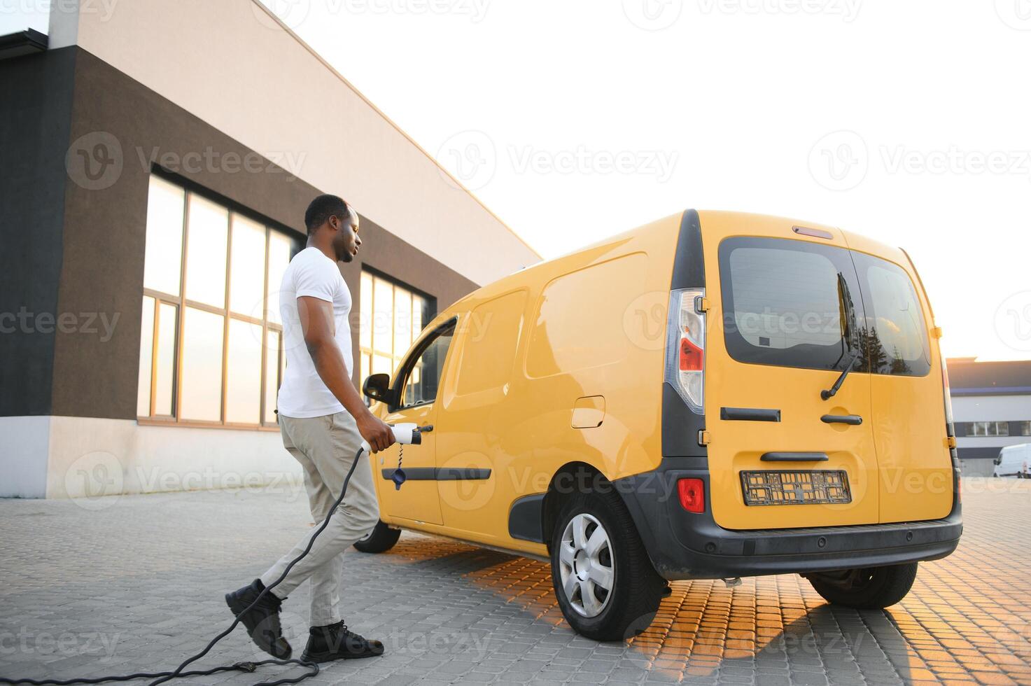 A african american man stands next to yellow electric delivery van at electric vehicle charging stations photo