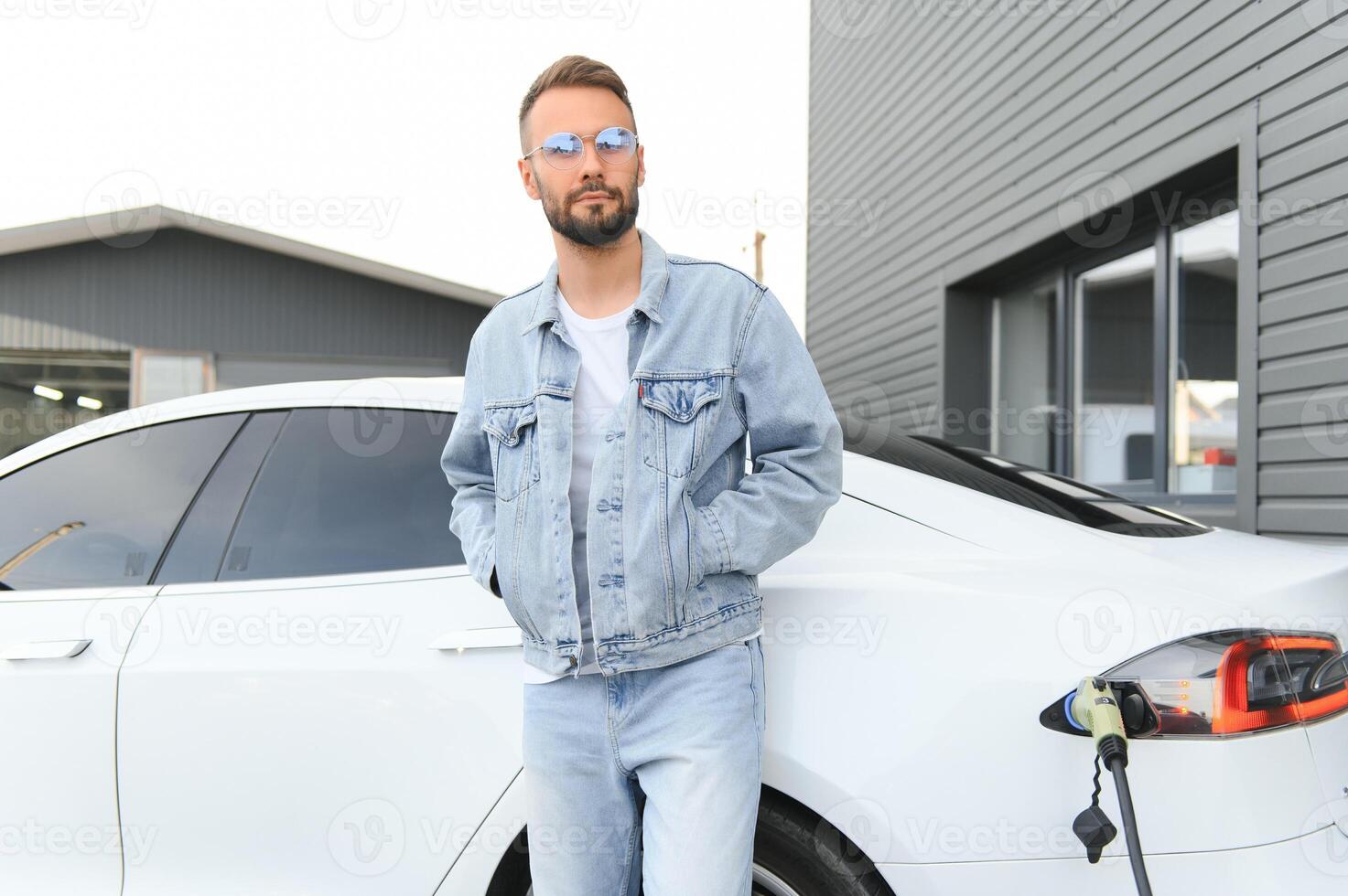 Standing and waiting for charge to finish. Young stylish man is with electric car at daytime photo