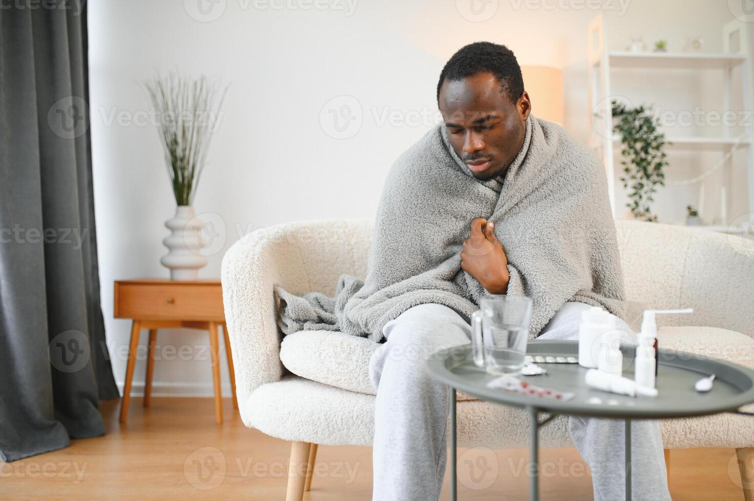 Cold african american young man covered in grey blanket freezing at home, sitting on sofa alone and warming up his hands in living room, copy space photo