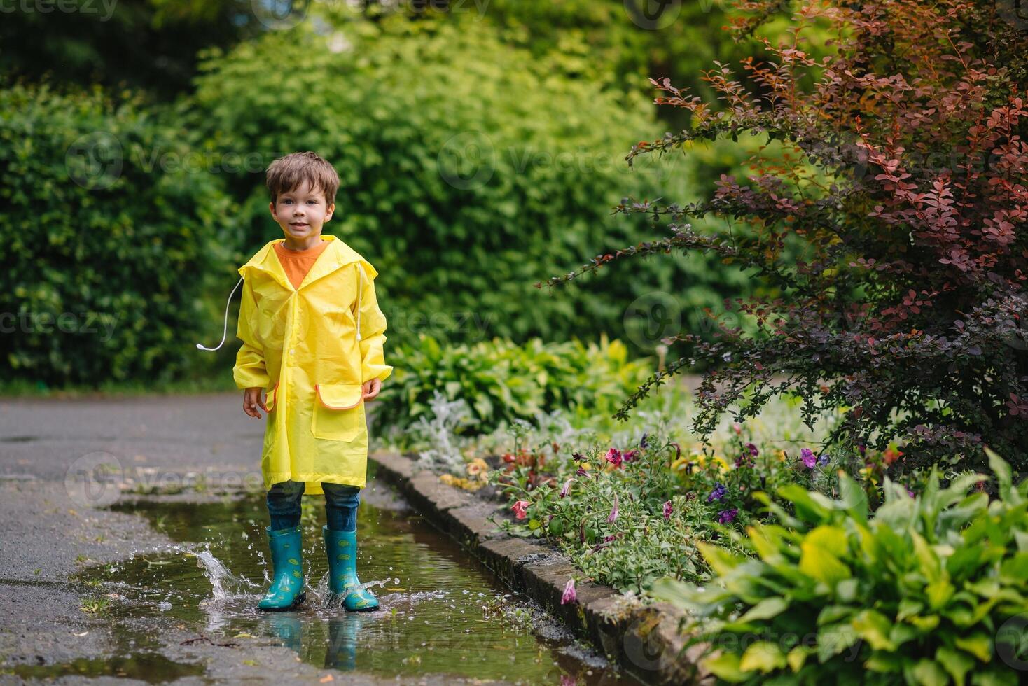 Little boy playing in rainy summer park. Child with umbrella, waterproof coat and boots jumping in puddle and mud in the rain. Kid walking in summer rain Outdoor fun by any weather. happy childhood. photo