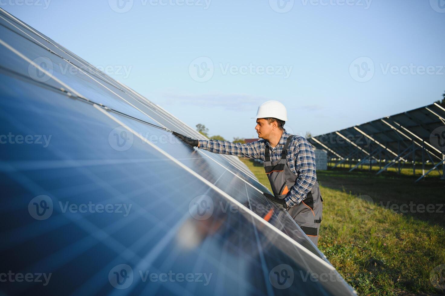 Portrait of Young indian man technician wearing white hard hat standing near solar panels against blue sky. Industrial worker solar system installation photo