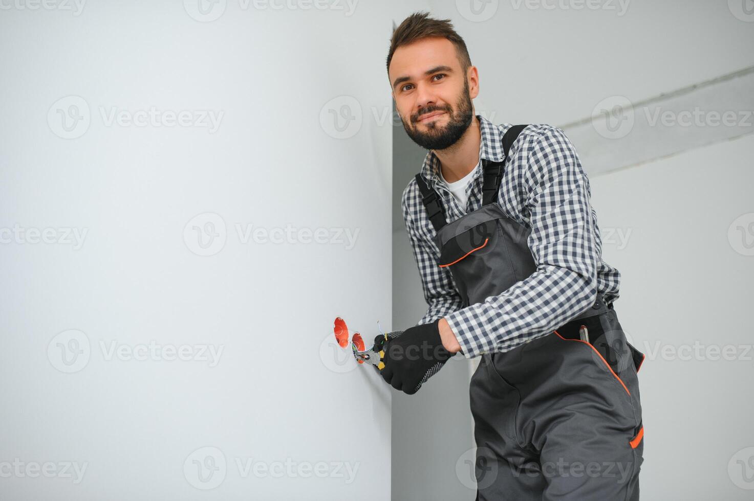 Electrician in uniform mounting electric sockets on the white wall indoors photo