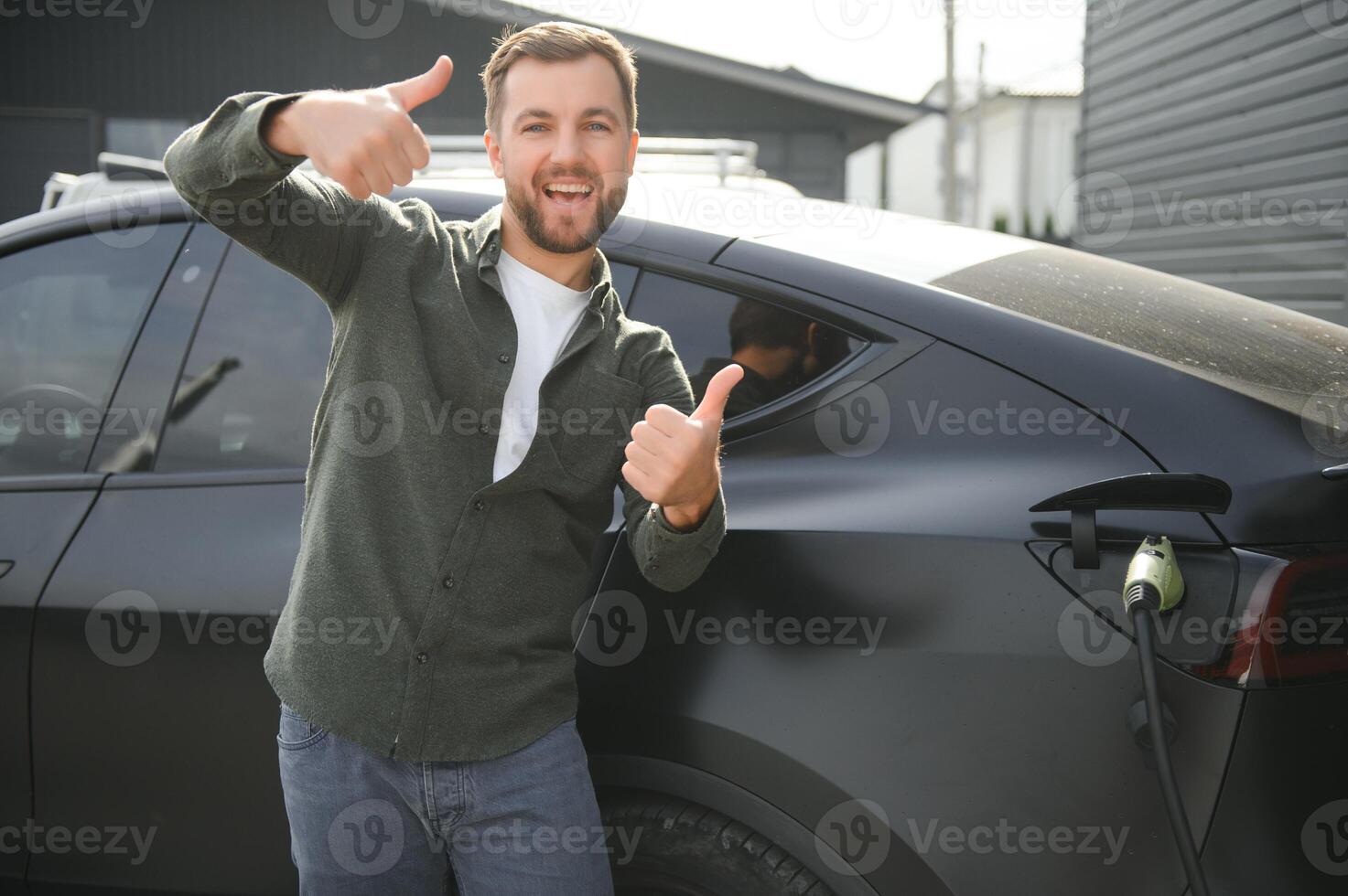 handsome bearded man in casual wear, standing at the charging station and charger for an electric car. Eco electric car concept photo