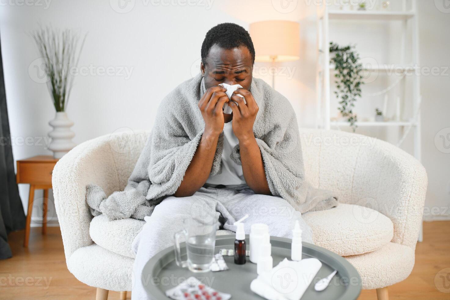 Portrait of a sick young african american man wrapped in a blanket on the sofa. Guy with seasonal flu or cold feel unhealthy with influenza and runny nose at home photo