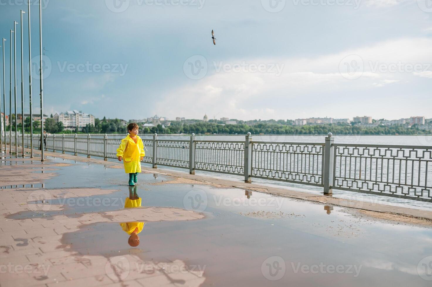 Little boy playing in rainy summer park. Child with umbrella, waterproof coat and boots jumping in puddle and mud in the rain. Kid walking in summer rain Outdoor fun by any weather. happy childhood. photo