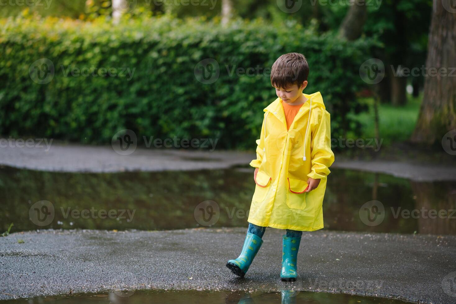 Little boy playing in rainy summer park. Child with umbrella, waterproof coat and boots jumping in puddle and mud in the rain. Kid walking in summer rain Outdoor fun by any weather. happy childhood photo