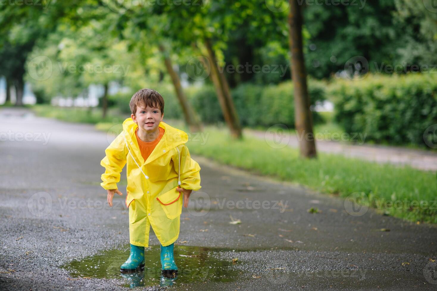 Little boy playing in rainy summer park. Child with umbrella, waterproof coat and boots jumping in puddle and mud in the rain. Kid walking in summer rain Outdoor fun by any weather. happy childhood photo