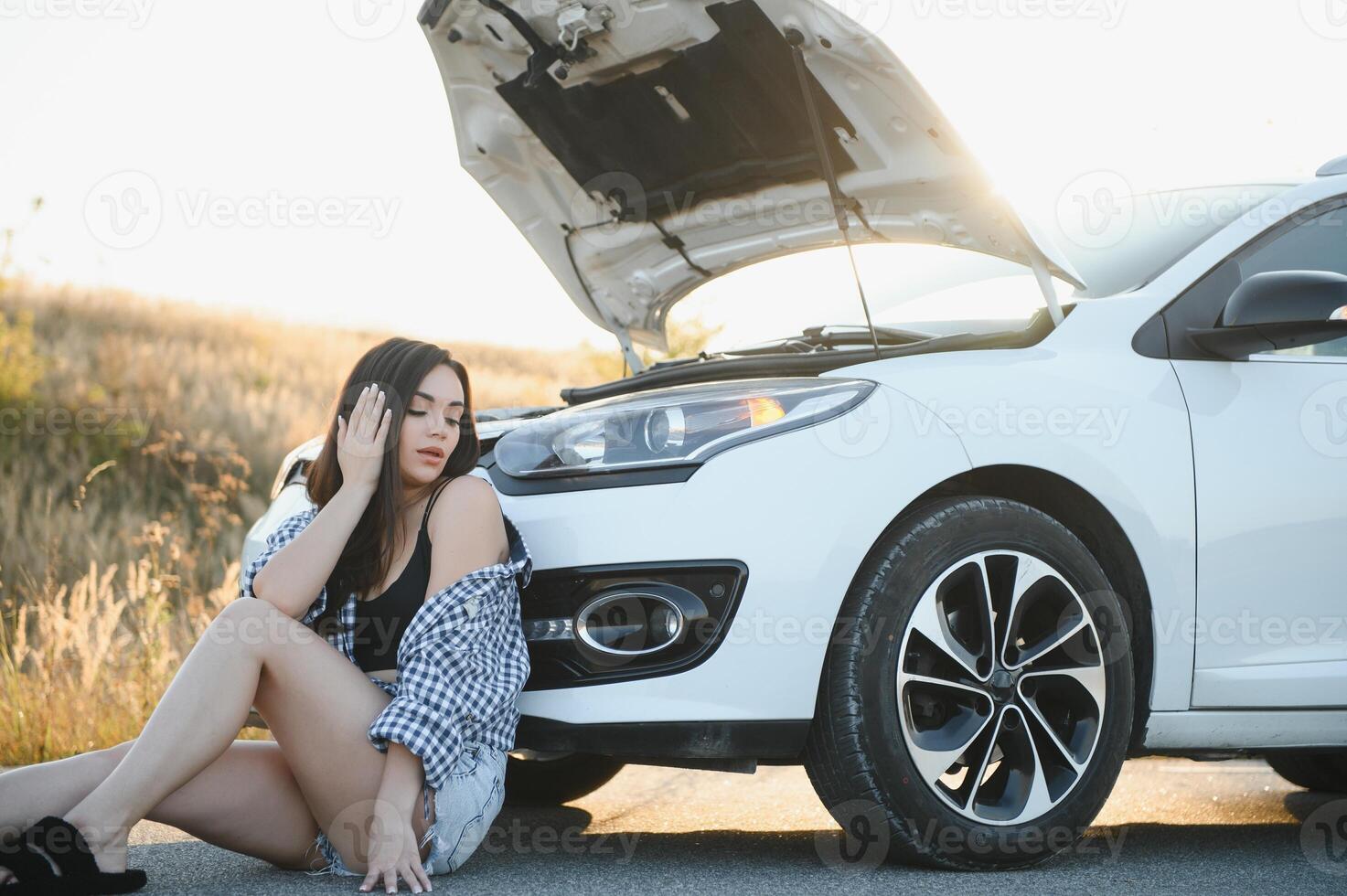 A young girl sits near a broken car on the road with an open hood. photo