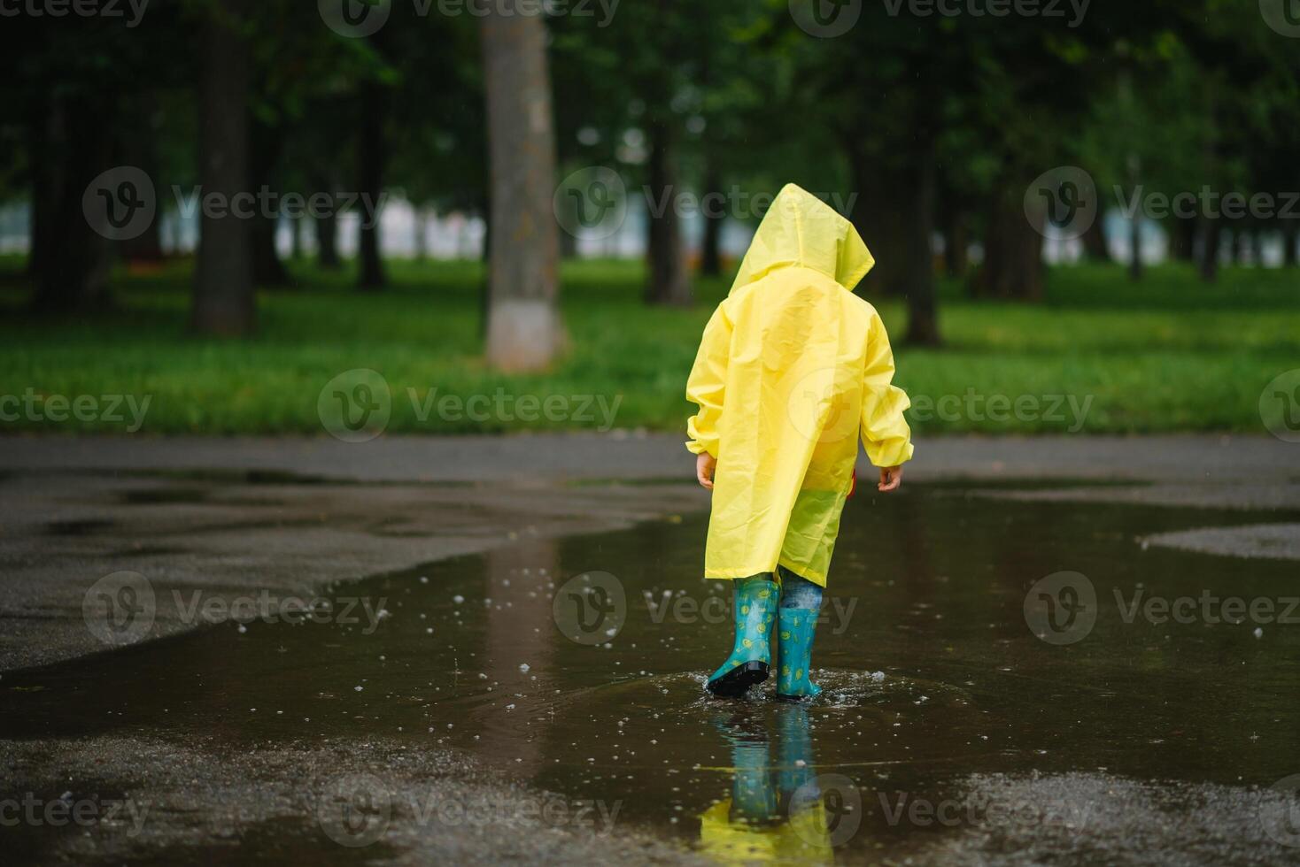 Little boy playing in rainy summer park. Child with umbrella, waterproof coat and boots jumping in puddle and mud in the rain. Kid walking in summer rain Outdoor fun by any weather. happy childhood. photo