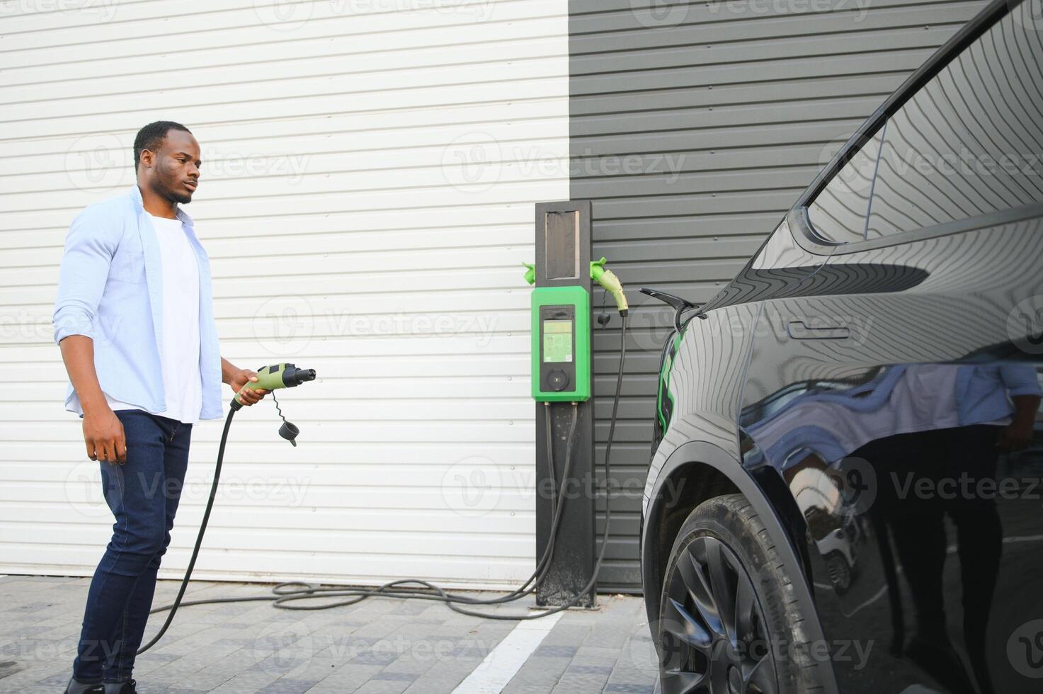 Serious african man holding charge cable in on hand, standing near luxury electric car. photo