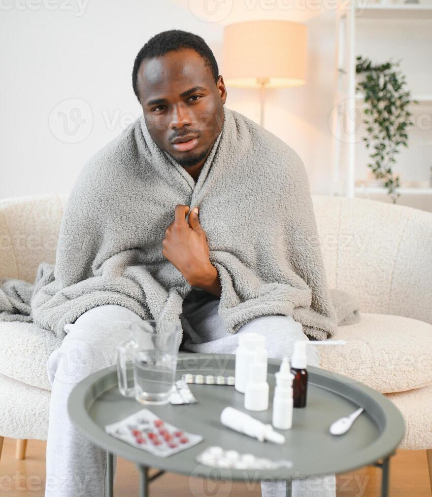 Cold african american young man covered in grey blanket freezing at home, sitting on sofa alone and warming up his hands in living room, copy space photo