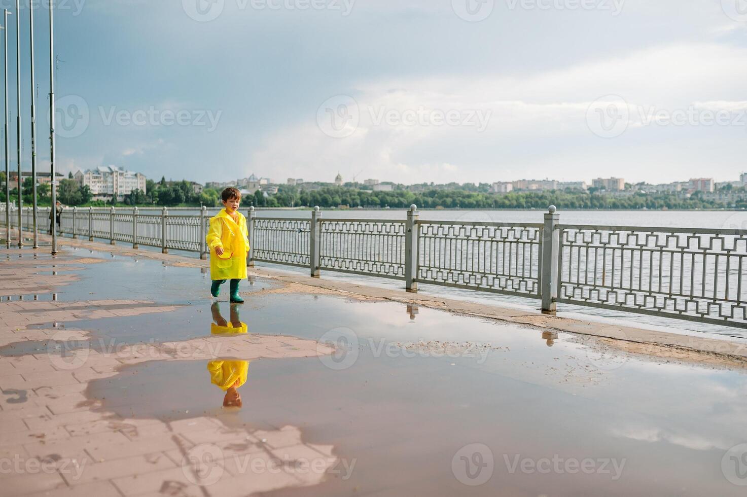 Little boy playing in rainy summer park. Child with umbrella, waterproof coat and boots jumping in puddle and mud in the rain. Kid walking in summer rain Outdoor fun by any weather. happy childhood. photo