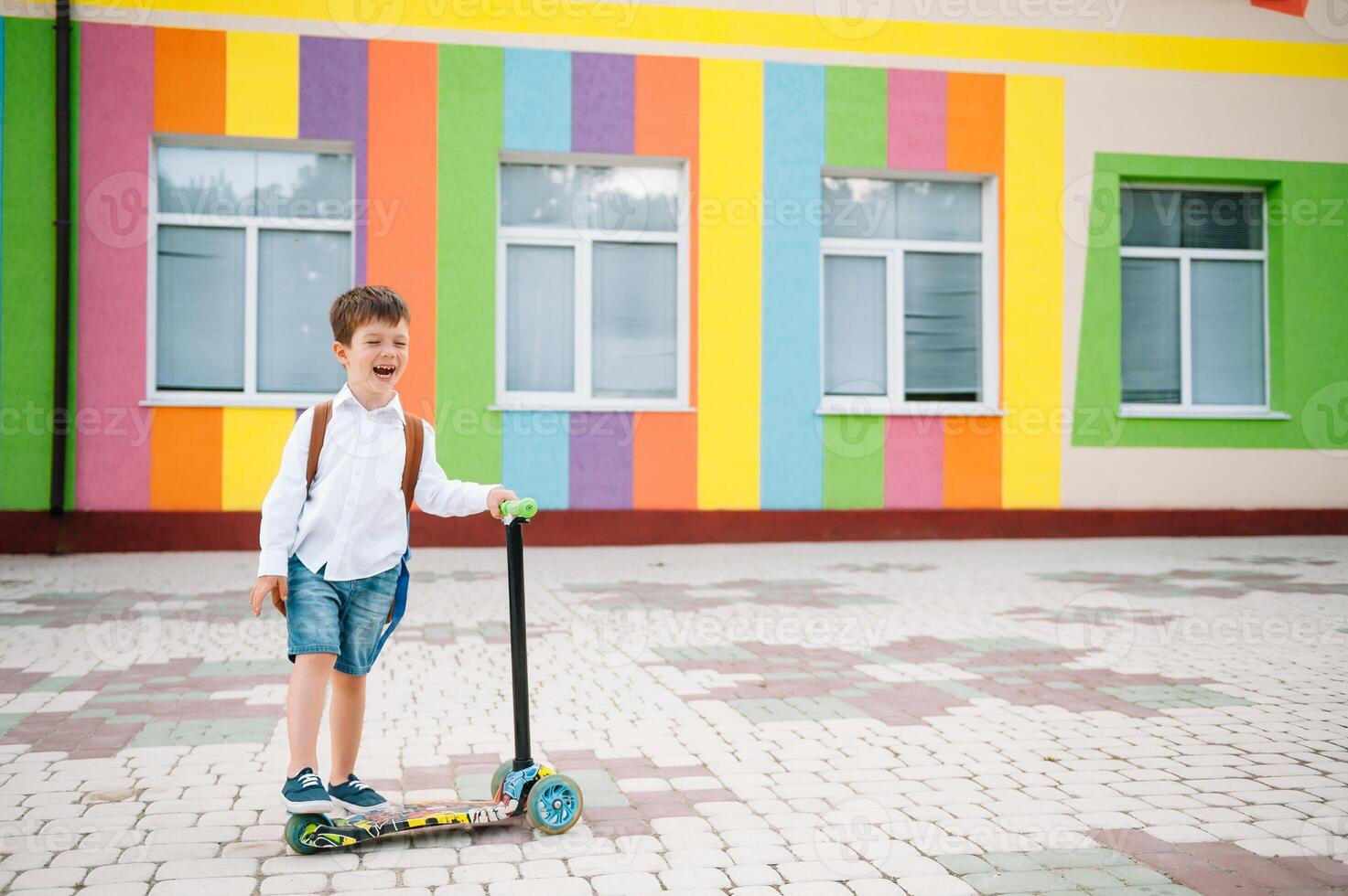 Adolescente chico con patada scooter cerca moderno escuela. niño con mochila y libro al aire libre. comenzando de lecciones primero día de caer. espalda a escuela. foto