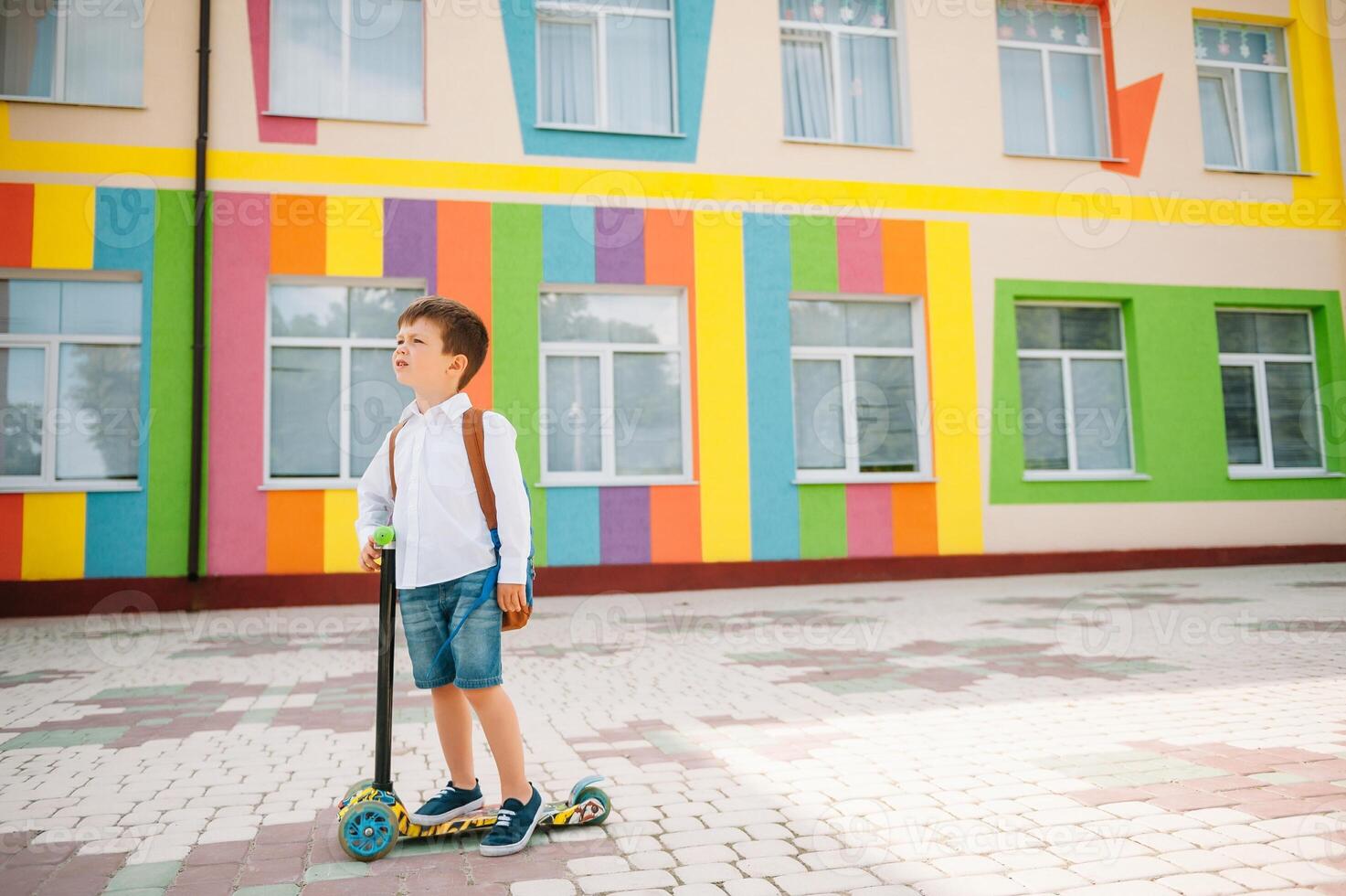 Teenage boy with kick scooter near modern school. Child with backpack and book outdoors. Beginning of lessons. First day of fall. Back to school. photo