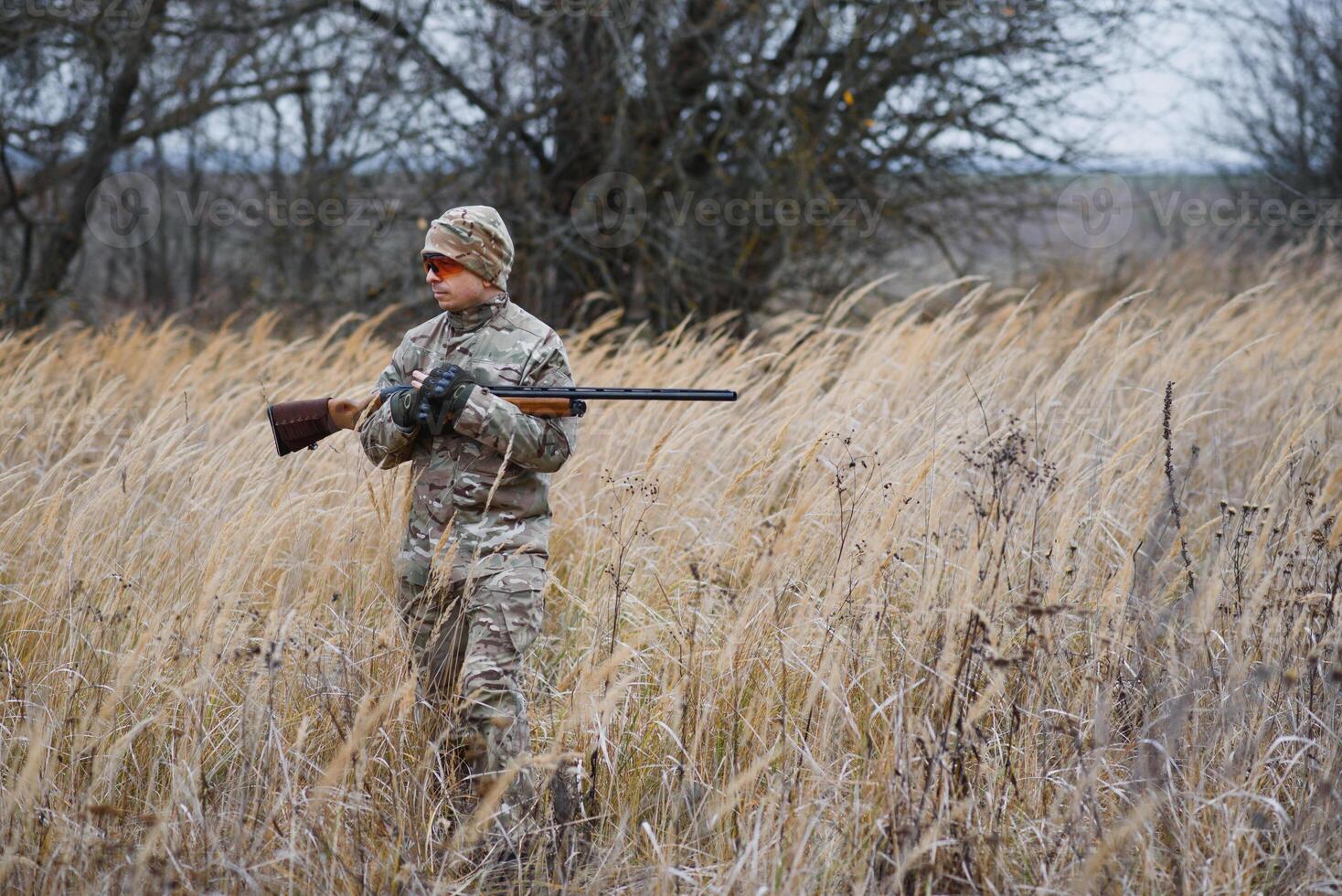 A male hunter with a gun while sitting takes aim at a forest. The concept of a successful hunt, an experienced hunter. Hunting the autumn season. The hunter has a rifle and a hunting uniform photo