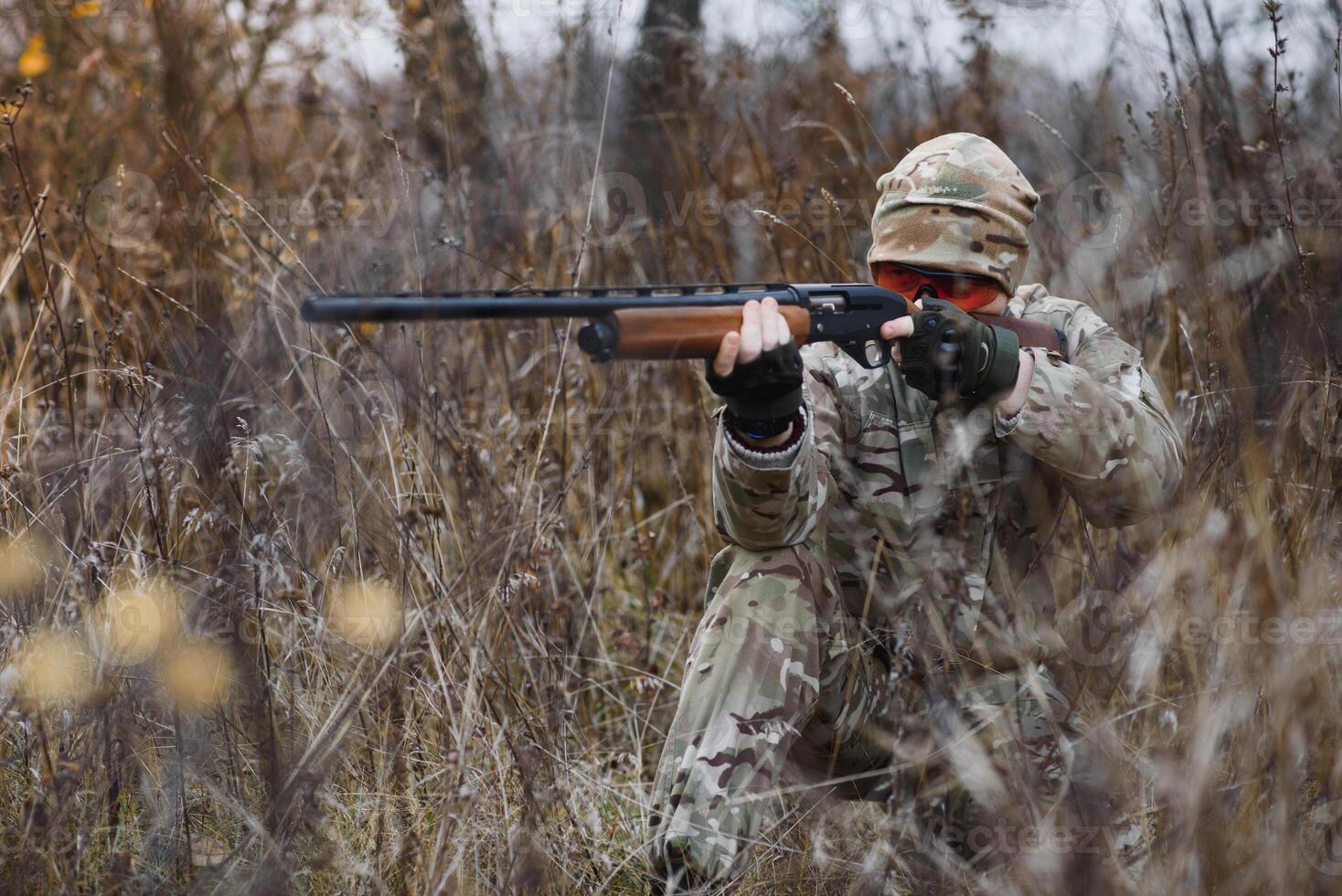 A male hunter with a gun while sitting takes aim at a forest. The concept of a successful hunt, an experienced hunter. Hunting the autumn season. The hunter has a rifle and a hunting uniform photo