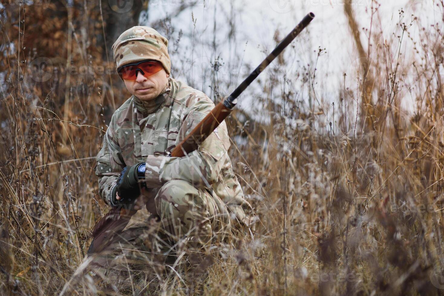 A male hunter with a gun while sitting takes aim at a forest. The concept of a successful hunt, an experienced hunter. Hunting the autumn season. The hunter has a rifle and a hunting uniform photo