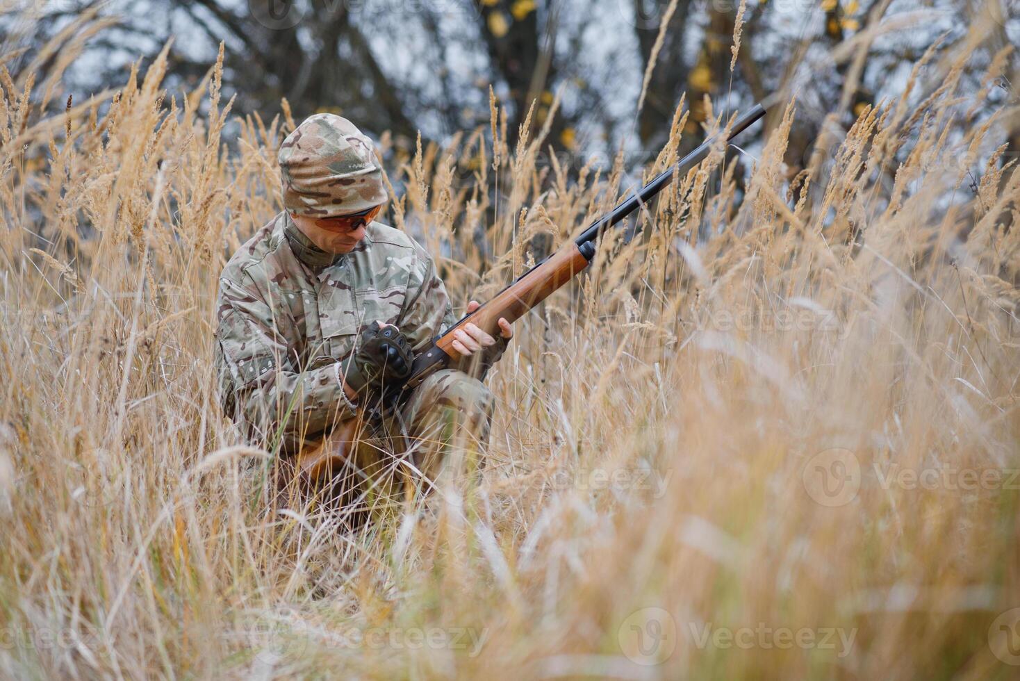 A male hunter with a gun while sitting takes aim at a forest. The concept of a successful hunt, an experienced hunter. Hunting the autumn season. The hunter has a rifle and a hunting uniform photo