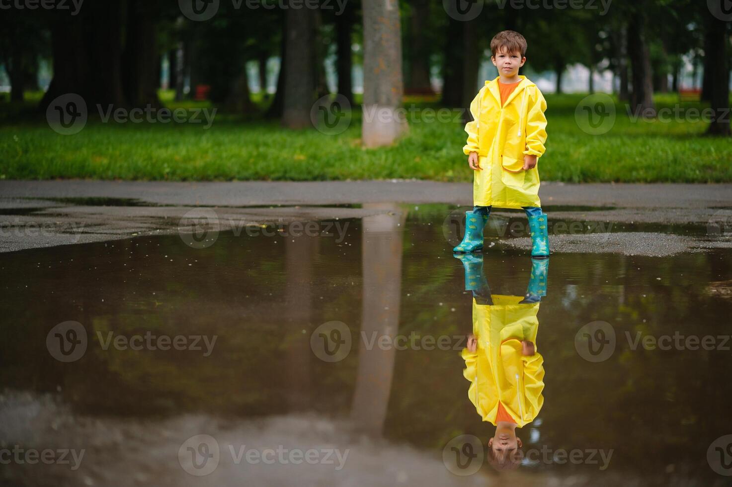 Little boy playing in rainy summer park. Child with umbrella, waterproof coat and boots jumping in puddle and mud in the rain. Kid walking in summer rain Outdoor fun by any weather. happy childhood. photo