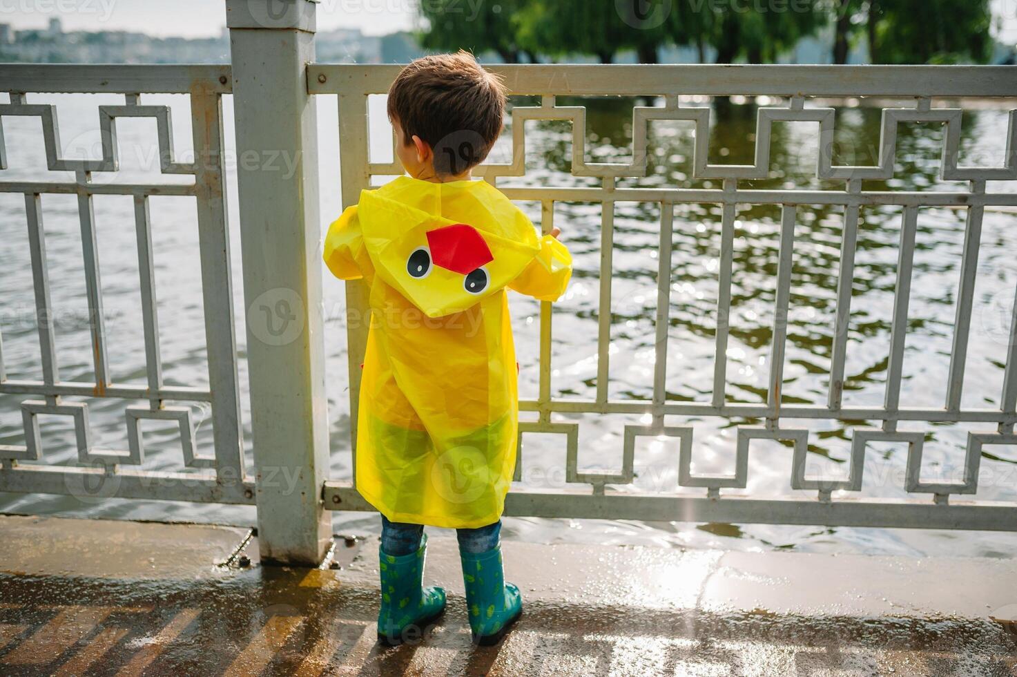 Little boy playing in rainy summer park. Child with umbrella, waterproof coat and boots jumping in puddle and mud in the rain. Kid walking in summer rain Outdoor fun by any weather. happy childhood. photo