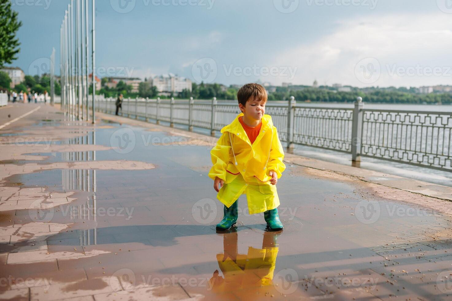 Little boy playing in rainy summer park. Child with umbrella, waterproof coat and boots jumping in puddle and mud in the rain. Kid walking in summer rain Outdoor fun by any weather. happy childhood. photo