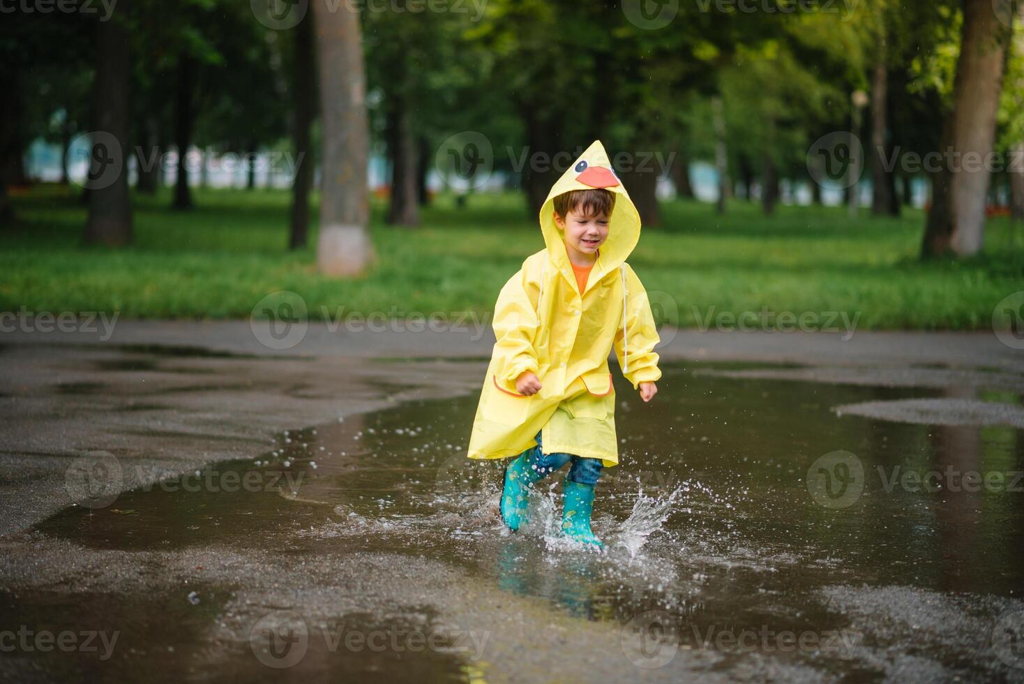 Little boy playing in rainy summer park. Child with umbrella, waterproof coat and boots jumping in puddle and mud in the rain. Kid walking in summer rain Outdoor fun by any weather. happy childhood. photo