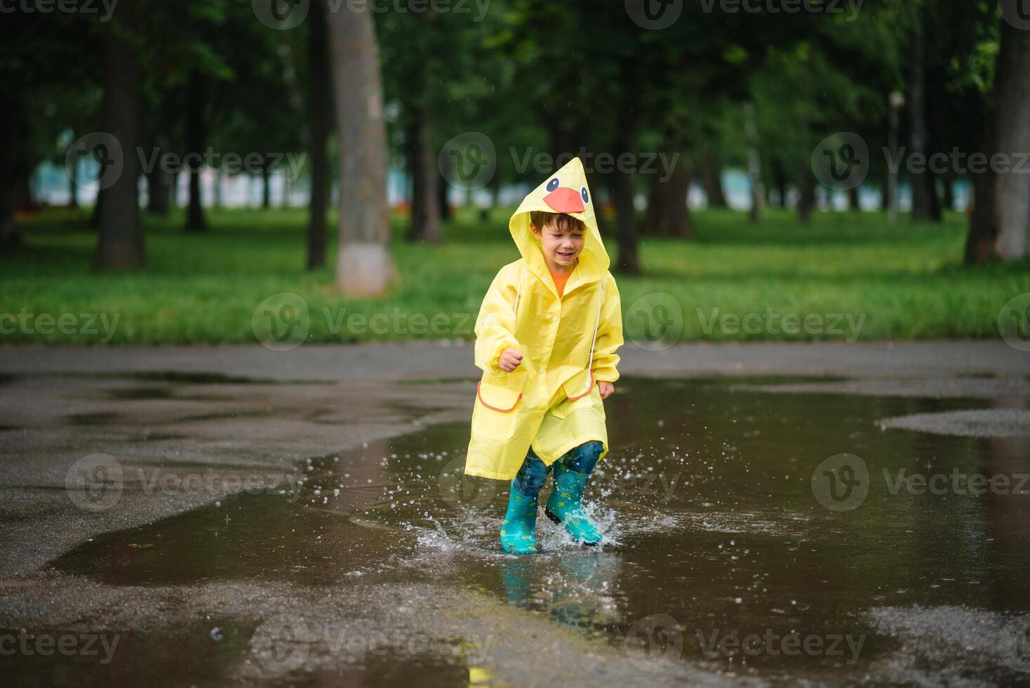Little boy playing in rainy summer park. Child with umbrella, waterproof coat and boots jumping in puddle and mud in the rain. Kid walking in summer rain Outdoor fun by any weather. happy childhood. photo