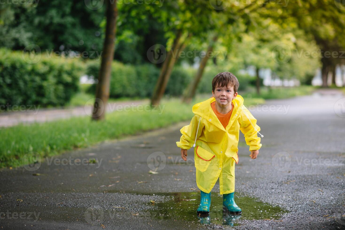 Little boy playing in rainy summer park. Child with umbrella, waterproof coat and boots jumping in puddle and mud in the rain. Kid walking in summer rain Outdoor fun by any weather. happy childhood photo