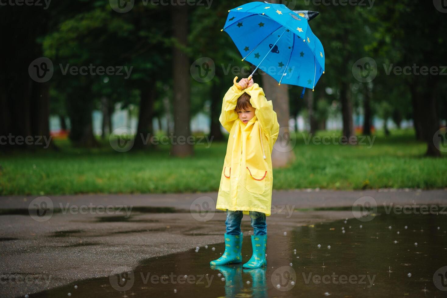 Little boy playing in rainy summer park. Child with umbrella, waterproof coat and boots jumping in puddle and mud in the rain. Kid walking in summer rain Outdoor fun by any weather. happy childhood. photo