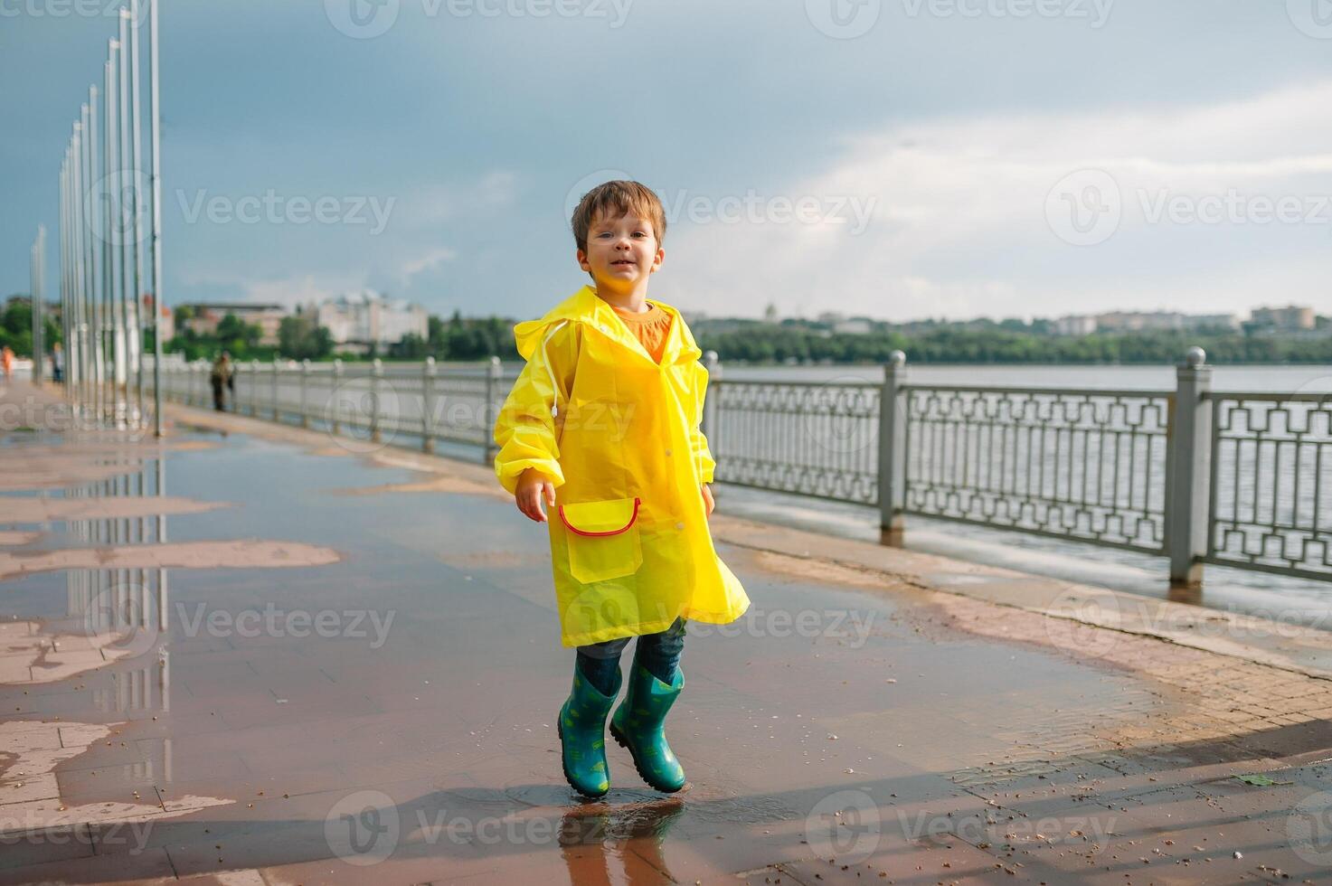 Little boy playing in rainy summer park. Child with umbrella, waterproof coat and boots jumping in puddle and mud in the rain. Kid walking in summer rain Outdoor fun by any weather. happy childhood. photo