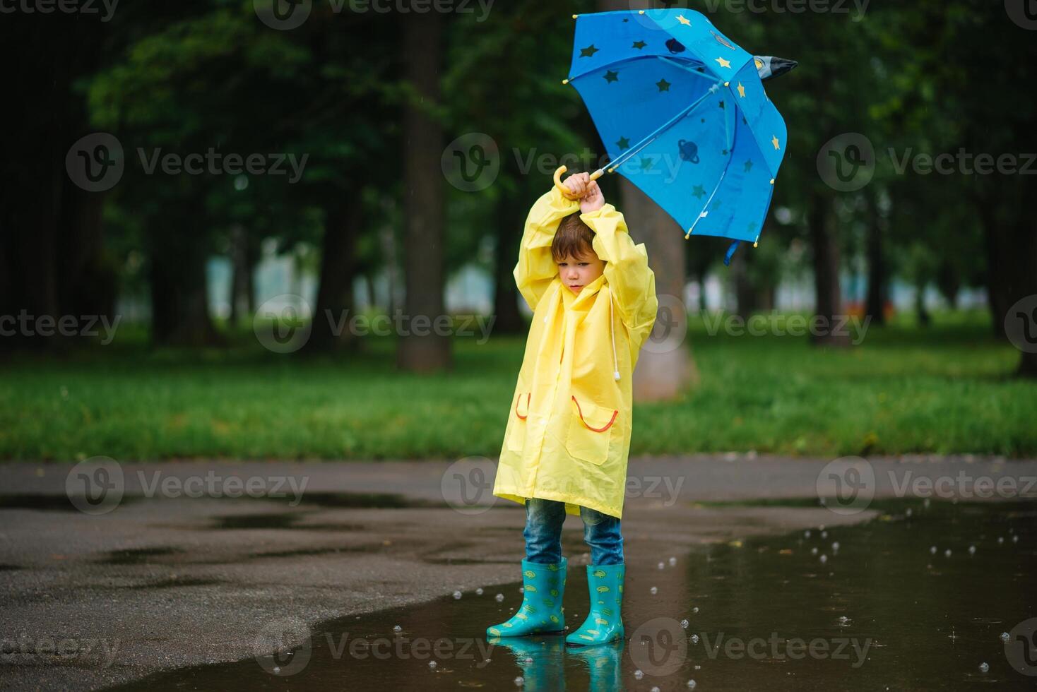 Little boy playing in rainy summer park. Child with umbrella, waterproof coat and boots jumping in puddle and mud in the rain. Kid walking in summer rain Outdoor fun by any weather. happy childhood. photo